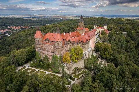 Zamek o czerwonych dachach wyrasta ze skalistego zbocza ponad lasem.