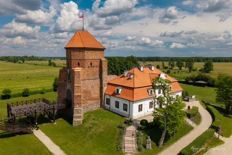 Ceglana wieża i czerwone dachy zamku otoczonego zielenią oraz otwartymi polami.