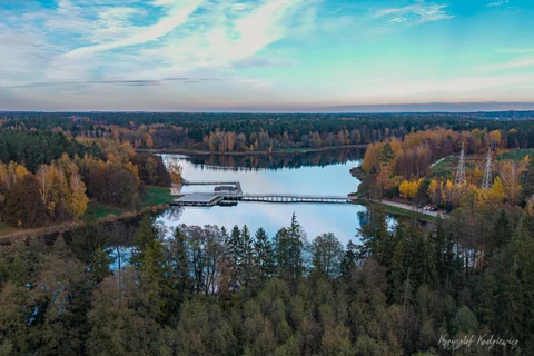 Pomost nad spokojną wodą otaczają jesienne drzewa i rozległy las pod pastelowym niebem.