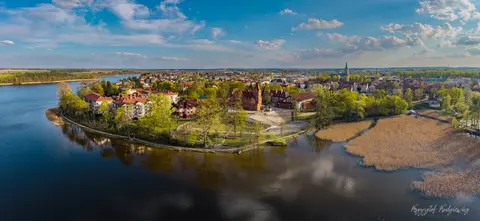 Zabudowa nadrzecznego miasta i zielone brzegi odbijają się w szerokiej tafli wody.
