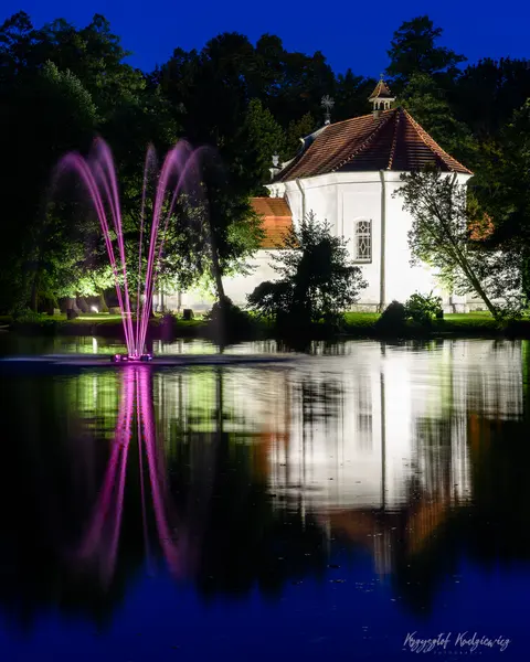 Oświetlona fontanna i jasny pawilon odbijają się w ciemnej tafli nocnego stawu.