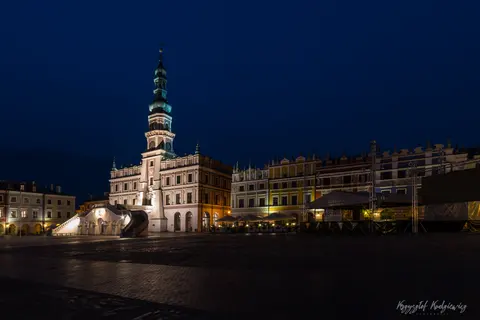Podświetlony ratusz i arkadowe kamienice otaczają rozległy plac po zmroku.