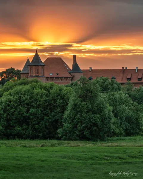 Czerwone dachy i wieżyczki ponad zielenią pod pomarańczowym pasmem zachodu.