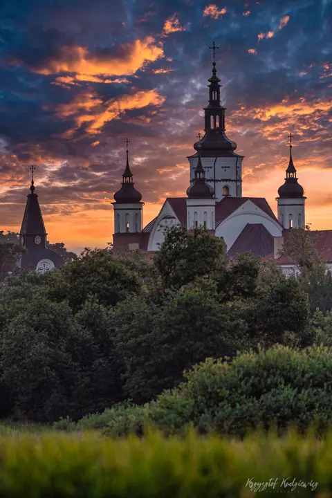 Wieże zabytkowej świątyni wyłaniają się znad drzew na tle barwnych chmur.