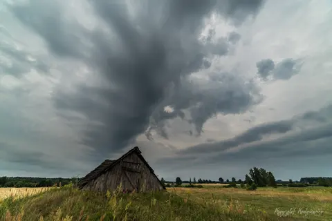 Ciężkie, poskręcane chmury zbierają się nad starą drewnianą chatą pośród pól.