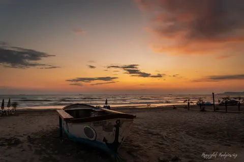 Łódź na pustoszejącej plaży pod pomarańczowymi chmurami nad spokojnym morzem.