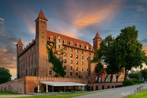 Wysoka ceglana wieża i rząd zamkowych okien w miękkim świetle wieczoru.