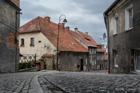 Brukowana uliczka zakręca między starymi domami o czerwonych dachach.