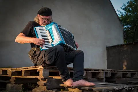Bosonogi akordeonista siedzi na drewnianych paletach z błękitnym instrumentem na kolanach.