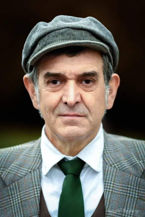 Wyrazisty portret mężczyzny w kaszkiecie, kraciastej marynarce i zielonym krawacie.