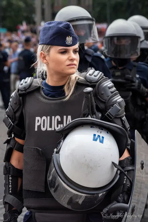 Policjantka z hełmem pod ramieniem na tle funkcjonariuszy w wyposażeniu ochronnym.