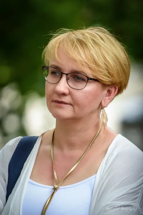 Kobieta w okularach i jasnym stroju, uchwycona w spokojnym portrecie plenerowym.