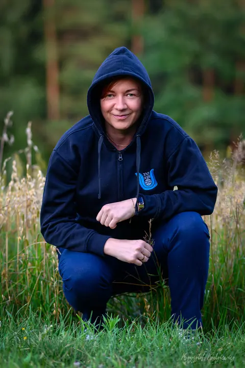 Mężczyzna w granatowej bluzie z kapturem przysiada wśród leśnych traw.