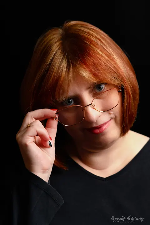 Rudowłosa kobieta spogląda znad opuszczonych okularów na głęboko ciemnym tle.