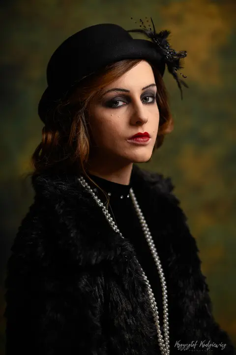 Portret w stylu retro: czarny kapelusz, długie perły i spojrzenie spod ronda.