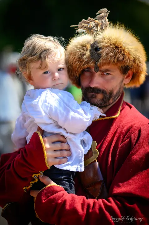 Mężczyzna w czerwonym, historyzującym stroju trzyma dziecko w jasnym ubraniu.