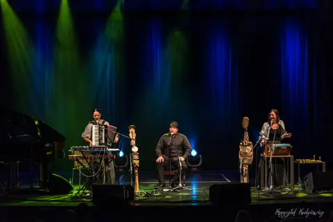 Artyści z akordeonem i instrumentami strunowymi na scenie w niebiesko-zielonym świetle.