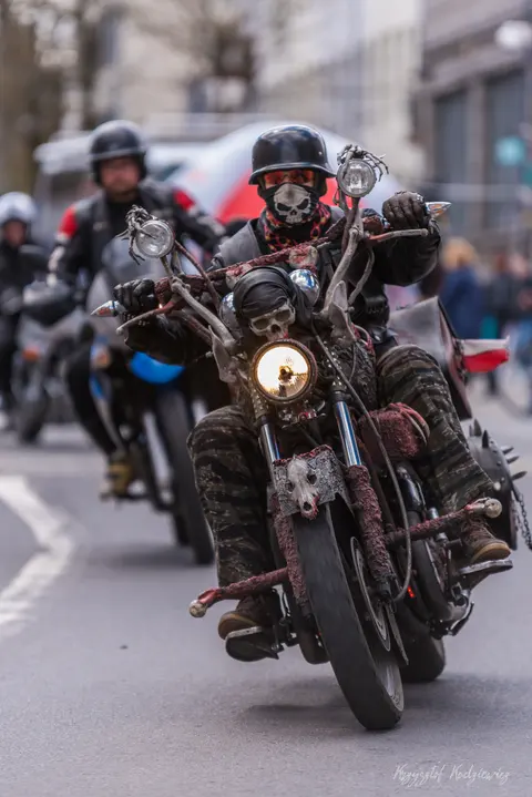 Motocyklista z malowaną osłoną twarzy prowadzi kolumnę maszyn miejską ulicą.