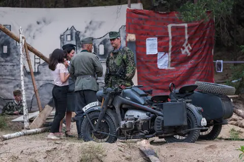 Motocykl z bocznym wózkiem i postacie w historycznych strojach przy scenografii budynku.