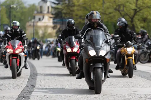Kolumna motocyklistów przejeżdża brukowaną ulicą w otoczeniu zielonych drzew.
