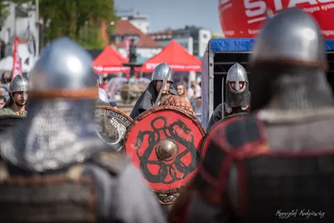 Czerwone tarcze widać pomiędzy nieostrymi hełmami stojących na pierwszym planie wojowników.