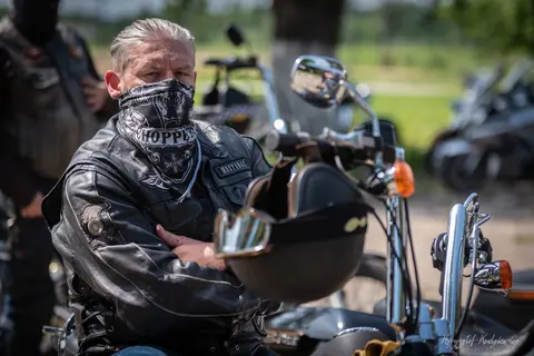 Motocyklista w skórzanej kurtce i chuście na twarzy siedzi za szeroką kierownicą.