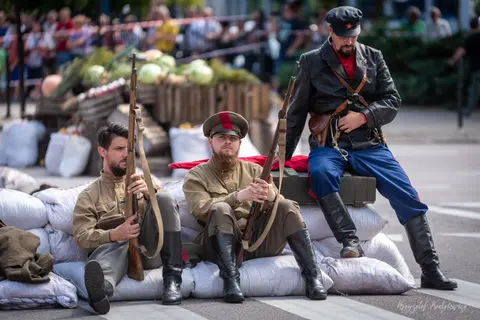 Postacie w historycznych strojach z długą bronią przy barykadzie z worków.