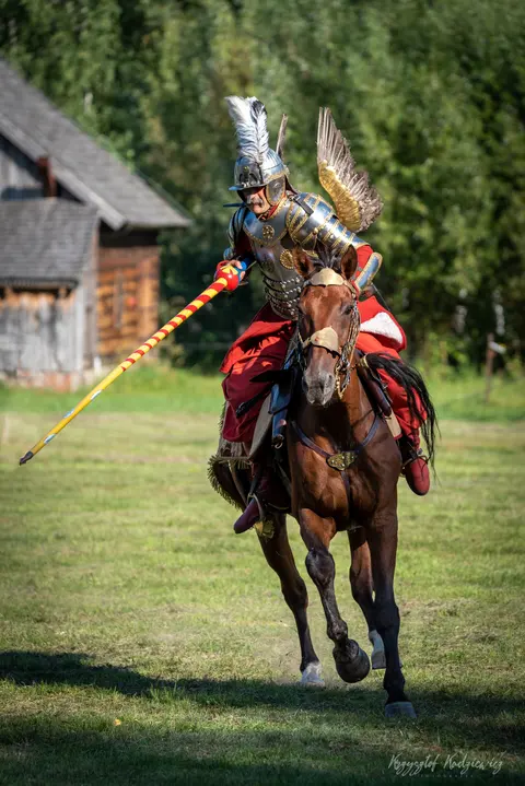 Jeździec w skrzydlatej zbroi prowadzi konia z kopią wysuniętą do przodu.