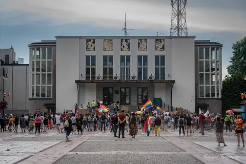 Tłum z tęczowymi flagami gromadzi się na placu przed monumentalnym budynkiem.