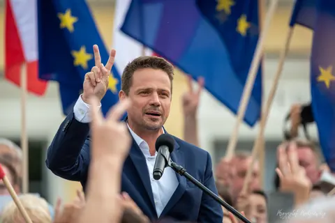 Mężczyzna przy mikrofonie unosi dłoń w geście zwycięstwa na tle flag.