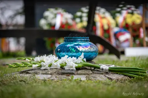 Niebieski znicz i białe kwiaty na kamieniu, z nagrobkami rozmytymi w tle.