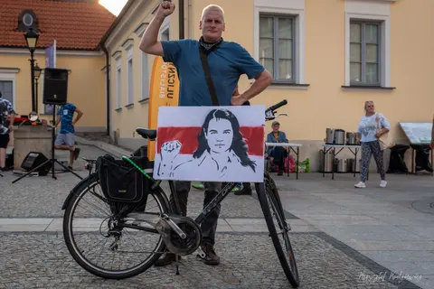 Mężczyzna przy rowerze unosi pięść nad tablicą z czarno-białym portretem.
