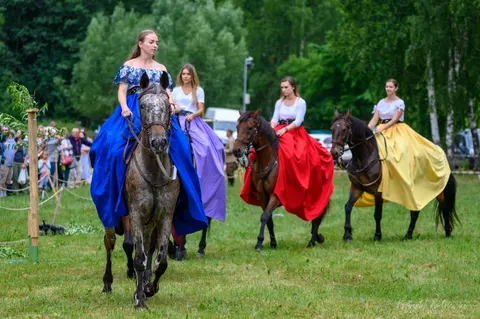 Kobiety w długich, barwnych sukniach jadą konno w równym szeregu po trawie.