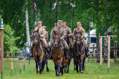 Trzej jeźdźcy w historycznych mundurach prowadzą konie, trzymając lance z proporczykami.