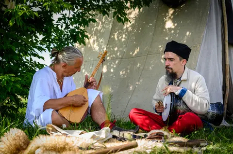 Muzycy z instrumentem strunowym i bębnem grają w cieniu plenerowego namiotu.