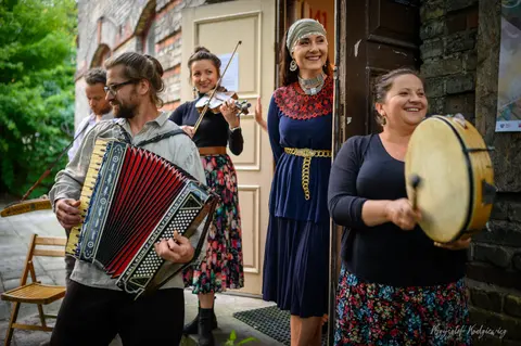 Akordeon, skrzypce i bęben towarzyszą wspólnemu muzykowaniu przed drewnianym budynkiem.