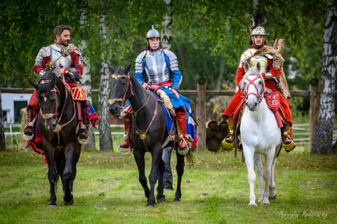 Trzej jeźdźcy w bogato zdobionych, historycznych strojach pozują na koniach.