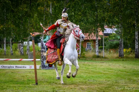 Jeździec ze skrzydłami i kopią prowadzi białego konia uniesionego w dynamicznym ruchu.