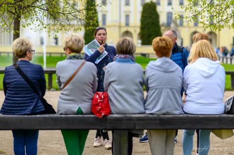 Grupa słuchaczy na parkowej ławce spogląda na kobietę przemawiającą przed pałacem.