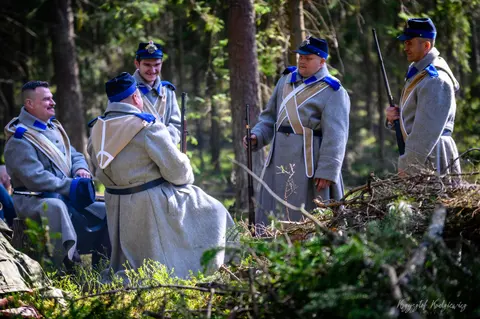 Rekonstruktorzy w szarych mundurach i niebieskich czapkach stoją pomiędzy leśnymi drzewami.